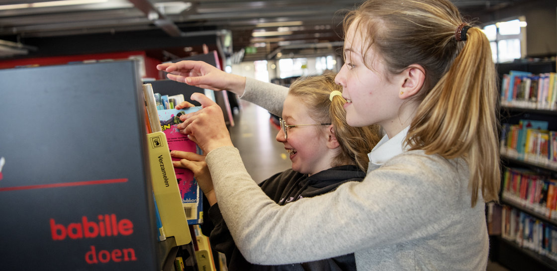 2 meisjes grijpen lachend naar een boek in het rek in de bib.