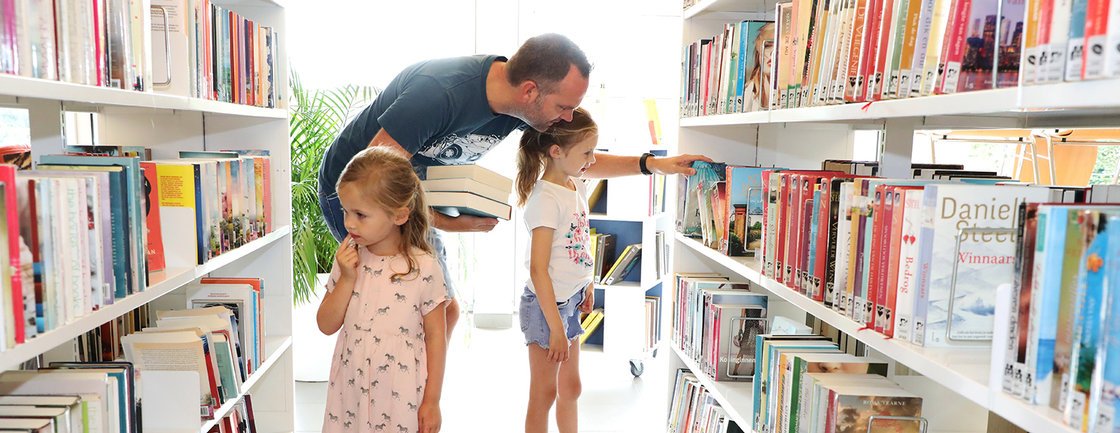 Man kiest boek met zijn dochters tussen de rekken van de bib.