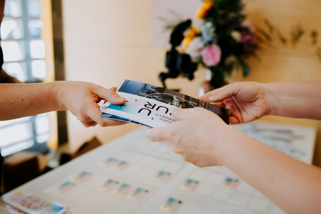 Een Cultuurconnect-collega geeft een folder van Café Connect futuur aan een bezoeker, met bloemen op de achtergrond