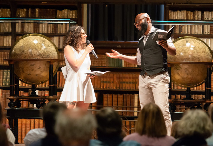 2 mensen op het podium, man en vrouw spreken tegen elkaar met wereldbol en boeken op de achtergrond
