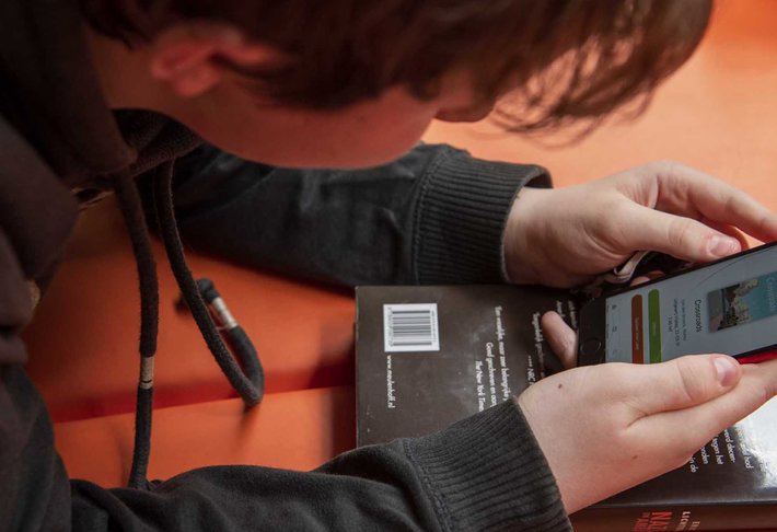 Jongere ligt neer en leest een e-boek, een fysiek boek ligt eronder.