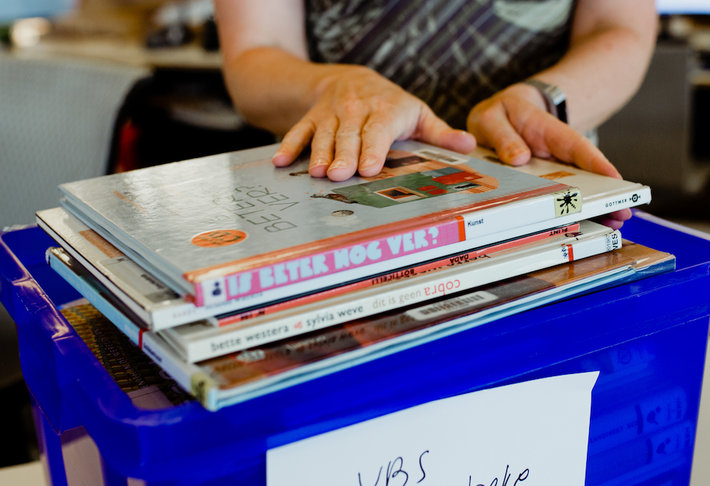 Een blauwe bak met boeken en 2 handen die er boeken in leggen.