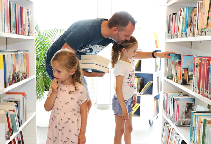 Man kiest boek met zijn dochters tussen de rekken van de bib.